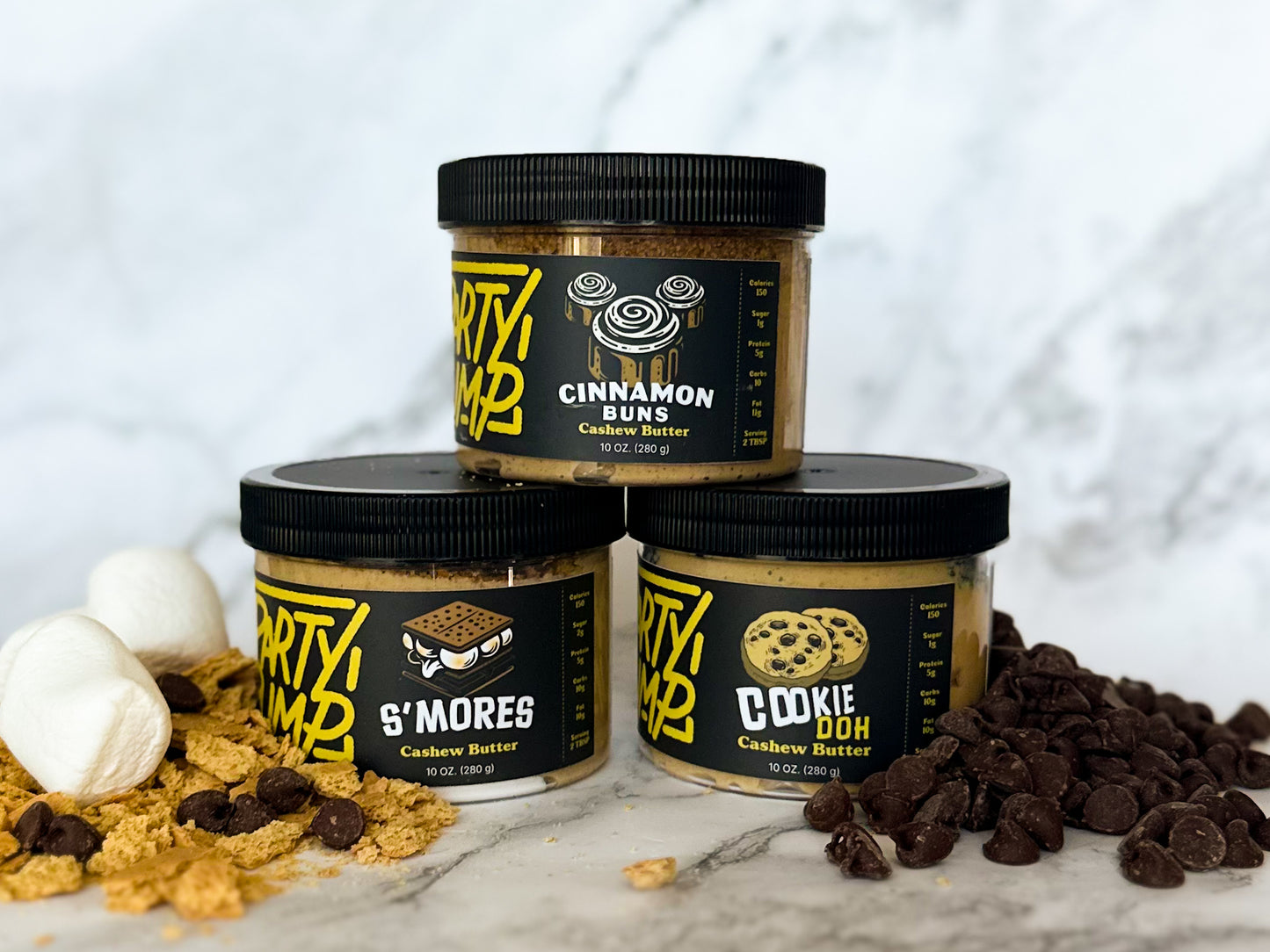 Three different flavors of cashew nut butter stacked in a pyramid. The flavors are as follows: Cinnamon Buns, Cookie Doh, and S'Mores. In the foreground there are ingredients representing the flavors.